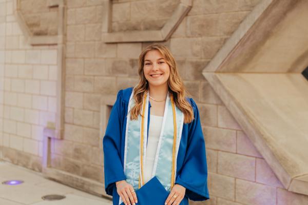 Ashley Grospitch wearing UK blue graduation gown, stoles, cords standing in front of the UK sign at the Gatton Student Center