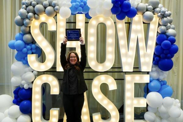 Dr. Martha Grady holding up an award in front of a lit sign spelling “Showcase” with blue and white balloons around it.