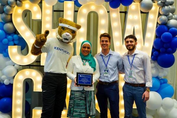 Yosra Helmy holds an award for Mentor of the Year standing by the Wildcat mascot and 2 of her students