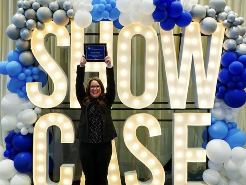 Dr. Martha Grady holding up an award in front of a lit sign spelling “Showcase” with blue and white balloons around it.