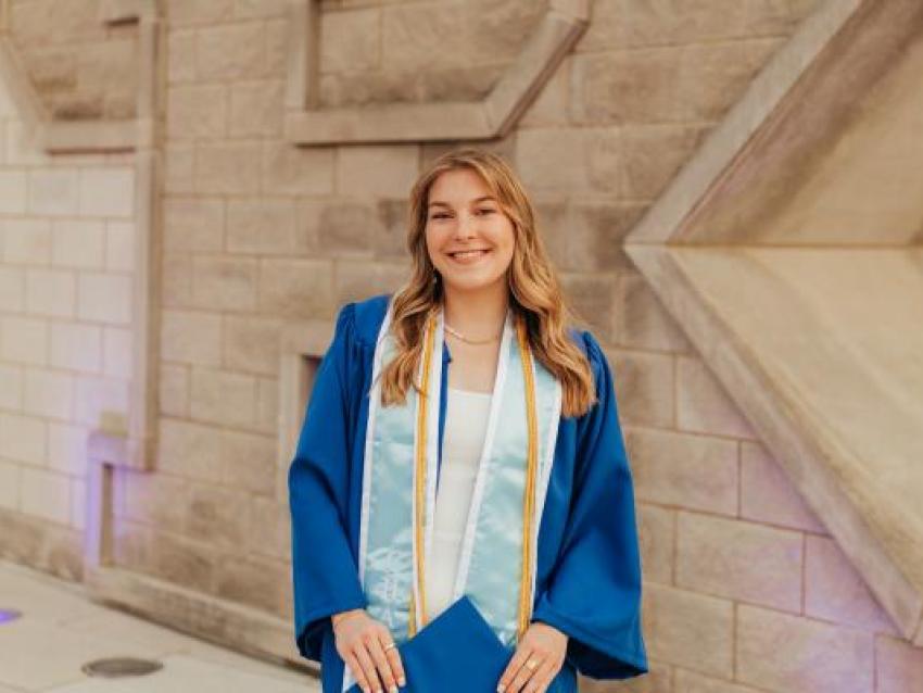 Ashley Grospitch wearing UK blue graduation gown, stoles, cords standing in front of the UK sign at the Gatton Student Center